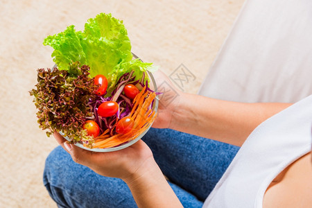 女手握碗腿上配有绿色生菜沙拉高于在碗里吃新鲜沙拉饭的年轻女在碗里吃素菜菠清洁去毒健康家庭自制食品概念图片