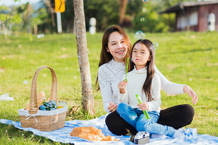 快乐的亚洲母亲和小女孩快乐的亚洲母亲和女儿玩得开心一起在户外享受快乐坐在草地上吹肥皂泡图片