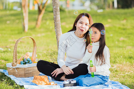 快乐的亚洲母亲和小女孩快乐的亚洲母亲和女儿玩得开心一起在户外享受快乐坐在草地上吹肥皂泡图片