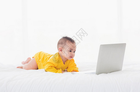 身着黄色礼服的亚洲小女孩在笔记本电脑键盘上亲手打字看白床上的监视器图片