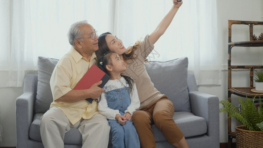 家庭快乐时间放松亚洲祖父母在家中用手机智能在沙发上与孙女一起笑着自拍老年男子或爷女儿和孩一起在客厅里图片