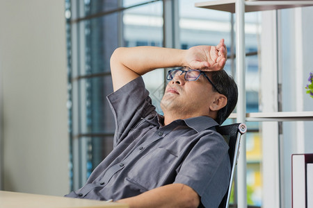 亚洲商人疲劳过度累他闭着眼睛放松双手在桌子上头顶着部身戴眼镜的老年人睡在工作场所的桌子上睡觉背景图片