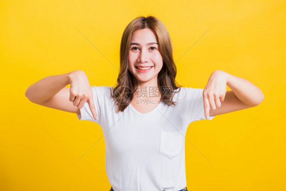 站在T恤衫上着的亚洲美丽可爱年轻女子手势在下面两根指点看着摄影机棚拍在黄色背景上图片