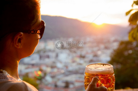 奥地利格拉茨市一位年轻女子坐在那里手里拿着一杯啤酒看着夕阳从Schlossberg山著名旅游景点钟楼的餐厅俯瞰奥地利格拉茨市一位图片