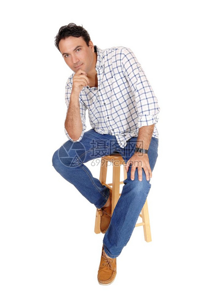 一个高的英俊男子坐在一个酒吧椅子上穿着一格衬衫和牛仔裤下巴上有一只手因白背景被隔离图片