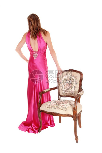 一个美丽的年轻女子穿着长的粉红色晚礼服站在后面的旧手椅旁与白背景隔绝图片