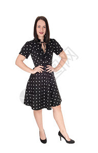 一个可爱的年轻女子站在从前高跟鞋和短黑头发穿着一副圆点的礼服与白背景隔绝图片