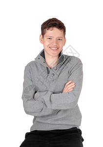 一个笑的年轻英俊少男孩坐在灰色的毛衣里双臂交叉与白种背景隔绝图片