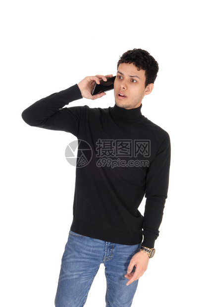 一个穿着黑色毛衣和牛仔裤站着的年轻瘦男子在电话上讲惊吓因白背景被孤立图片