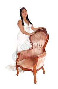 一位可爱的新娘穿着白色婚纱站在古老的董粉红色手椅后面笑着与白色背景隔绝图片