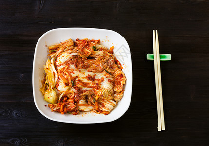 korean烹饪白碗中Kimchi开胃菜辣椒小麦卷心菜和黑棕色木制餐桌上筷子的顶端视图图片
