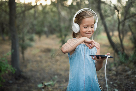 微笑金发女郎白色的小孩在森林里用她的智能手机和耳监听音乐图片