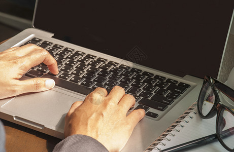 年轻的一位正在办公室的木头上打笔记本电脑并贴有笔和眼镜的年轻女子近视图像该少女在她的办公室用笔记本电脑打字一种套装图片