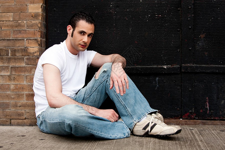 哈基又黑色的地面身着白衬衫和蓝牛仔裤身着侧角烧伤的白人英俊男子坐在黑金属车库门前水泥地板上的混凝土图片