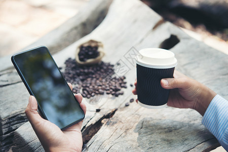 手持纸杯咖啡和智能手机的男子坐在人手上拿着纸杯咖啡和智能手机坐在木制桌子上的男带走休息早晨背景图片