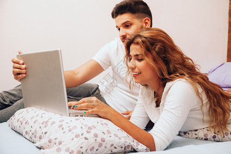 朋友们微笑着的年轻异恋夫妇躺在床上用笔记本电脑爱枕头图片
