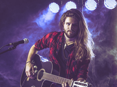 聚光灯演员音乐家照片来自一位长发头留胡子的年轻人在舞台上弹着声响吉他有灯光和音乐会气氛A图片