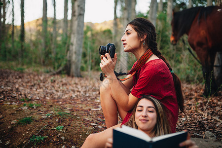 类比的小型黑发年轻笑着微的一对年轻女穿着阅读书本的裙子在森林里用旧相机拍照图片