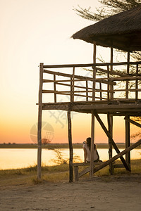 一名年轻女旅游客坐在一个破屋顶小下拍摄非洲博茨瓦纳MakgadikkadkadiPans的日落照片在下面人们放松图片