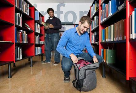 男人跪下从背包里拿东西在图书馆之间的公共图书馆里而另一个人则在背景中失去焦点笑声在室内架子大学图片