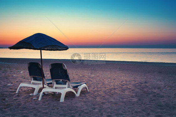 闲暇在日落的桑迪海滩沙上的伞状和两张阳床在海边和日落天空背景下带有防晒床的桑迪海滩放松和旅行的保护伞概念以及日落时在海滩上的两个图片