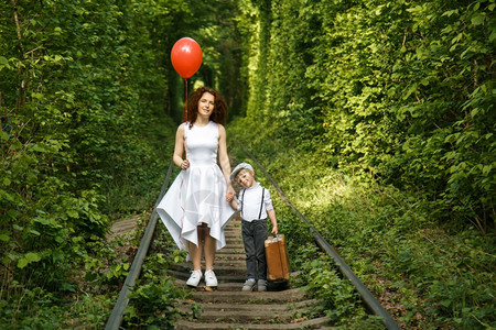 年轻的有红气球和小男孩的女人在森林隧道的铁路上大手提箱旅行概念走自然图片