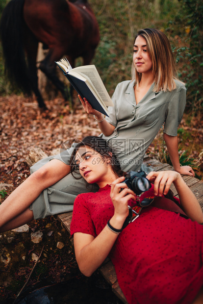 年轻的一对身着洋装的年轻女子正在阅读一本书森林里用旧相机和马拍照恶魔女图片