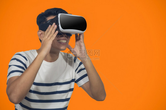 有虚拟现实头盔的年轻人站在橙色背景的室内青年拿着虚拟现实头盔的年轻人接触眼镜耳机图片