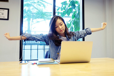松弛在办公桌上用笔记本电脑工作办公室生活方式商业状况时年轻的青女在从事个人电脑工作时伸展身体以放松力为了桌子图片