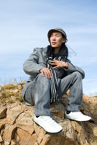 身着灰色大衣和条纹黑白围巾的年轻亚洲男子坐在岩石上他脸的痕迹是古蒙语中他的名字Chinghis黑色的亚洲人外套图片