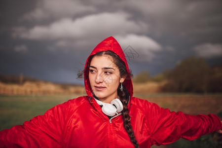 编织多云的年轻女子在雨天与衣和耳机一起站在田地上图片