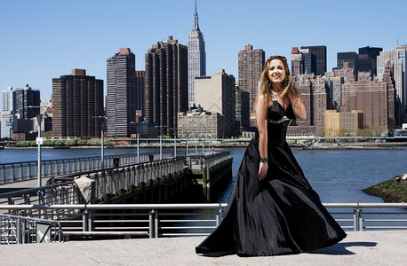 穿着黑晚鸡尾酒礼服的美丽快乐高加索拉丁时装模特女郎站在纽约市曼哈顿天际线前站着状态户外图片