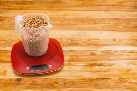 展示准备鹰嘴豆在透明量杯中天然木材背景上的红色塑料电子秤上称重图片