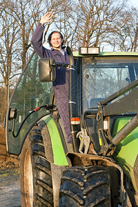 单身的一只快乐农手站在大拖拉机开着的门上对摄像机挥手泥肮脏的图片
