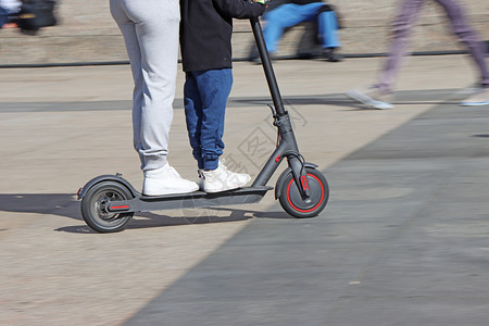 电动滑板车轮母亲和儿子在市广场乘坐电动脚踏车运模糊城市的背景