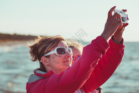 太阳镜女孩移动的暑假期间母亲和女儿在海上用智能手机照相拍摄片的背面景象图片