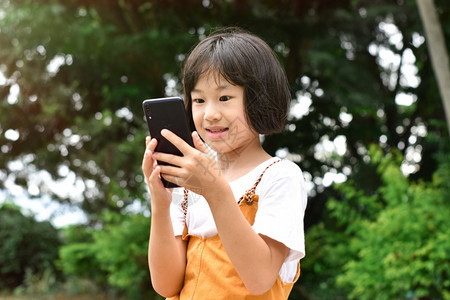 互联网可爱的电话在公园使用移动智能手机的快乐女孩Asian快乐儿童在公园里笑着看设备屏幕Kid正在微笑和观看通讯技术的概念图片