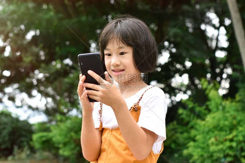 互联网可爱的电话在公园使用移动智能手机的快乐女孩Asian快乐儿童在公园里笑着看设备屏幕Kid正在微笑和观看通讯技术的概念图片