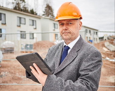 蓝色的欣德斯特伦成熟男工程师使用平板电脑他穿着西装和安全帽背景是房屋建筑工地图片