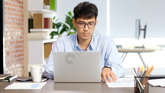 互联网考虑年轻的亚洲商人专注于办公室使用笔记本电脑手提笔记本电脑亚洲办公用人Comtemplate板亚洲男商人和办公室生活方式概图片