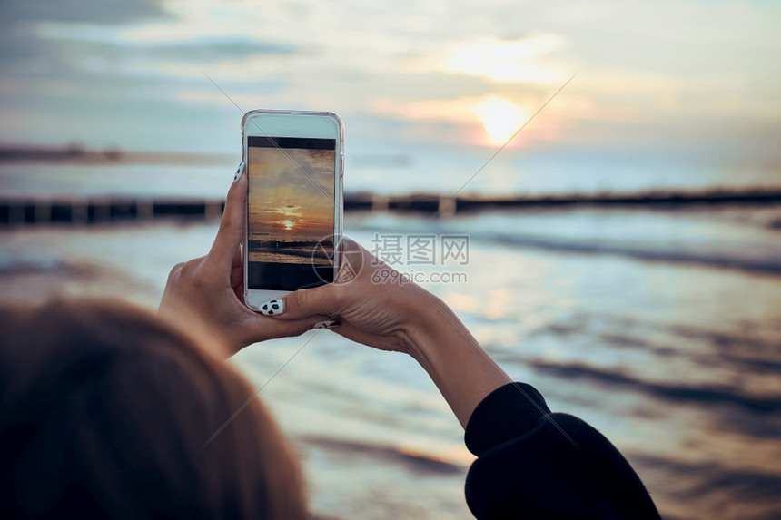 女年轻子在暑假旅行中使用智能手机拍摄海面日落的照片对女孩持有智能手机和拍照的视觉观移动背光图片