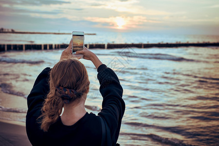 游客电话年轻女子在暑假旅行中使用智能手机拍摄海面日落的照片对女孩持有智能手机和拍照的视觉观移动图片