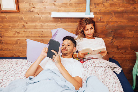 在室内松弛朋友们年轻笑着的异恋夫妇躺在床上看书用平板电脑写字图片