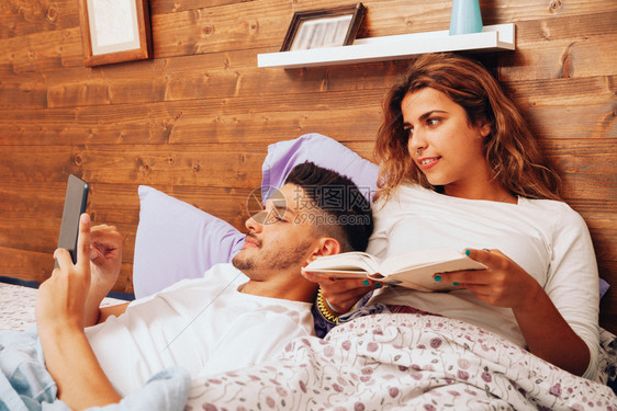 卧室年轻笑着的异恋夫妇躺在床上看书用平板电脑写字说谎木头图片