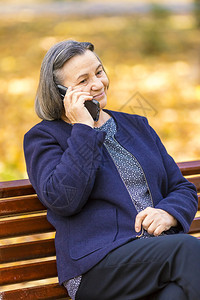 坐着请讲移动的正面高级女在公园户外使用智能手机以电话为重点图片