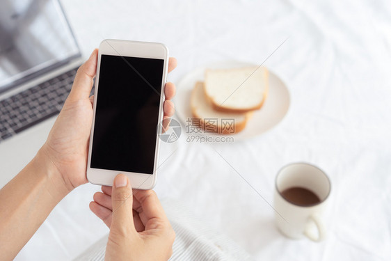 女士糕点保持工作妇女手握并用智能电话在办公桌背景上持有和使用智能手机的在桌上有咖啡面包和笔记本电脑图片