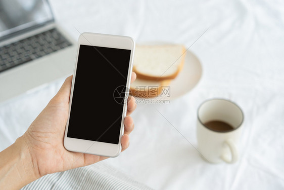 女聪明的工作妇女手握并用智能电话在办公桌背景上持有和使用智能手机的在桌上有咖啡面包和笔记本电脑打字图片