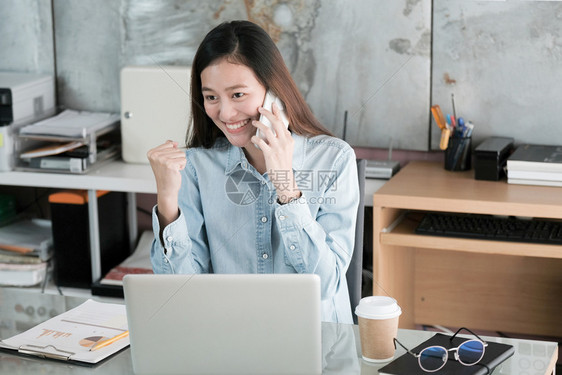 年轻的亚洲女商人在办公桌工作时用微笑的面脸说话讲电和手举起来商业概念取得成功在职的兴奋尽管图片