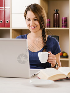 浏览在线的吸引人一个美丽微笑着女士使用笔记本电脑在家或办公室喝咖啡的照片图片