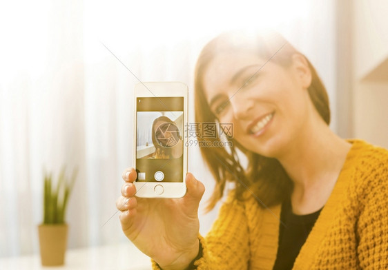 服用移动的有魅力女人在家里用智能手机拍自己照片时带着她的智能手机脸图片